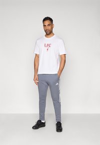 Λευκή κοντομάνικη μπλούζα με κόκκινο λογότυπο "LFC", συνδυασμένη με γκρι στενές φόρμες με λευκές πλευρικές ρίγες και μαύρα αθλητικά παπούτσια.