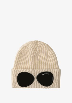 Gorro de punto color crema con textura acanalada, que presenta lentes negros parecidos a goggles en la parte frontal. Logo de la marca "C.P. COMPANY" visible en los lentes.