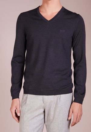 Mann trägt einen dunkelblauen V-Ausschnitt Pullover mit langen Ärmeln und hellgraue Hose vor einfachem Hintergrund.