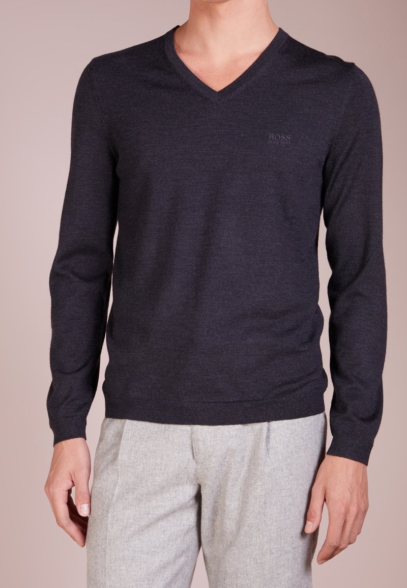 Homme portant un pull à col en V à manches longues de couleur bleu marine foncé et un pantalon gris clair, sur un fond uni.