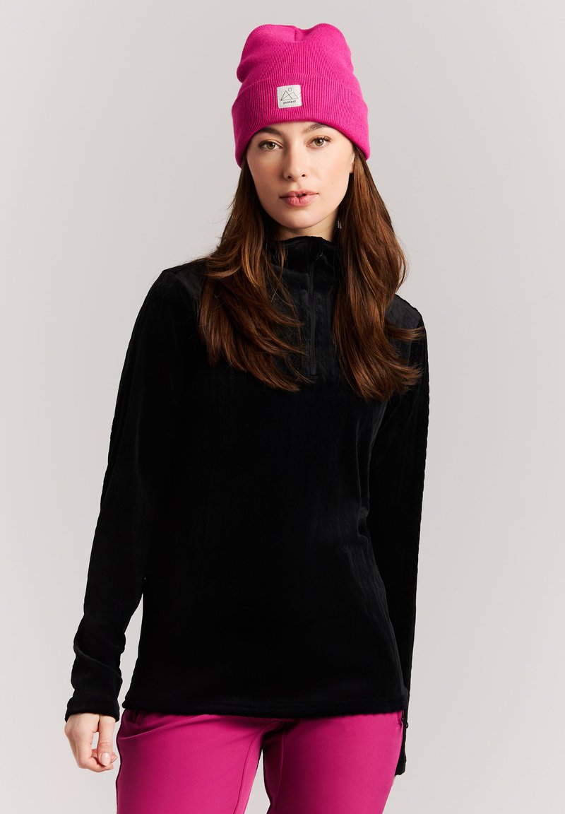 Jeune femme portant un bonnet rose vif, un pull noir à manches longues et un pantalon rose vif assorti sur un fond clair uni.