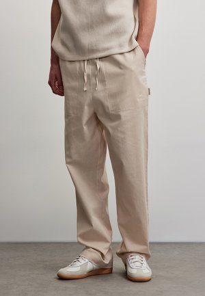 Persoon draagt beige broek met trekkoord en bijpassende top, witte en beige sneakers, staand op grijze vloer tegen een effen muur.