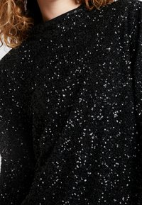 Svart paljetttopp med en strukturerad yta, med en oversized passform och rund halsringning, som reflekterar ljuset tack vare de många glänsande paljetterna.