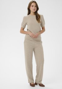 Beżowy sweter z krótkimi bufiastymi rękawami, połączony z pasującymi beżowymi spodniami o szerokich nogawkach. Buty są brązowe, plecione, na słupku. Gładka tekstura.