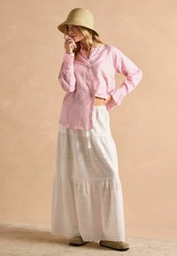Woman wearing a straw hat, pink striped button-up shirt, white eyelet maxi skirt, and beige shoes, standing against a beige background.