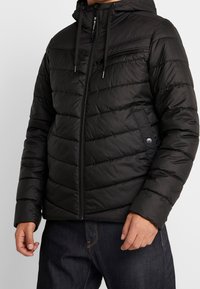 Svart puffjacka med dragkedja på framsidan, huva och quiltad design. Funktioner inkluderar en bröstficka och elastiska mudd. Slät textur.