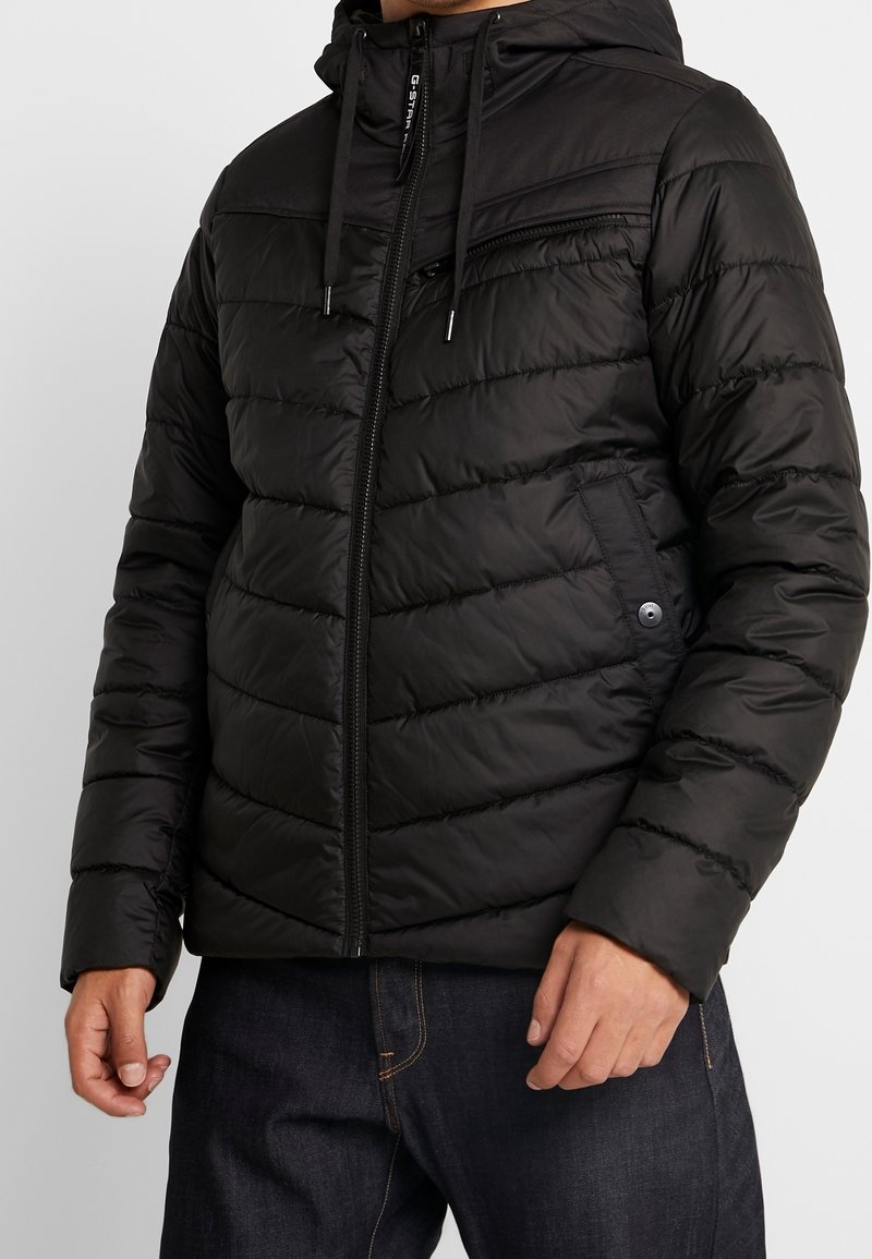 Svart puffjacka med dragkedja på framsidan, huva och quiltad design. Funktioner inkluderar en bröstficka och elastiska mudd. Slät textur.