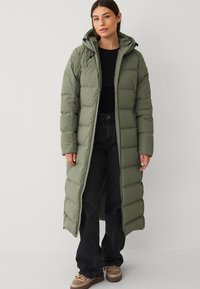 Lunga giacca imbottita verde oliva con cappuccio, caratterizzata da sezioni trapuntate e chiusura con zip frontale. Indossata sopra un top nero e jeans scuri.