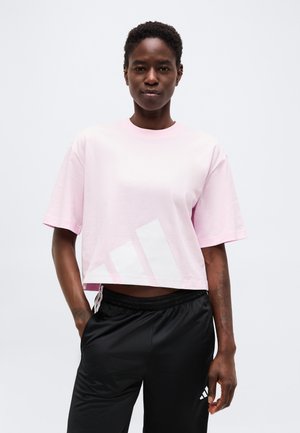 Frau trägt ein lockeres, hellrosa, kurzes T-Shirt mit weißem Logo und schwarze Sporthose mit weißen Streifen, steht mit einer Hand in der Tasche.