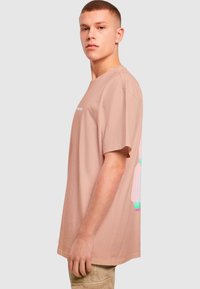 Světle růžové oversized tričko vyrobené z bavlny. Na zadní straně má barevný grafický design a kulatý výstřih. Krátké rukávy.