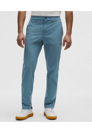 Mężczyzna noszący wąskie jasnoniebieskie spodnie i białe trampki z gumowymi podeszwami, stojący na tle gładkiego jasnego tła.