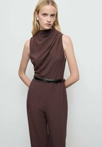 Bruine mouwloze jumpsuit met een hoge gedrapeerde hals en een aansluitende taille, geaccentueerd door een zwarte riem. Glad materiaal met een gestructureerde vorm.