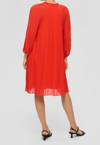 Rotes plissiertes Kleid mit langen Ärmeln, Rundhalsausschnitt und strukturiertem Stoff. Kombiniert mit schwarzen Sandalen mit Absatz, die das fließende Design des Kleides zur Geltung bringen.