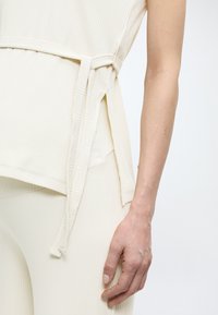 Detailní záběr na trup a paži osoby v žebrovaném krémovém topu a odpovídajících kalhotách, s postranním vázacím detailem na topu.