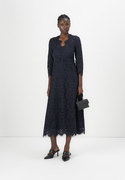 Navy lace dress with three-quarter sleeves, scalloped neckline, and hem. Accessorized with a small black handbag and black heeled mules.