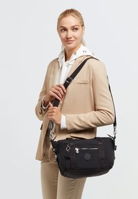 Black fabric shoulder bag with a smooth texture, featuring a silver zipper, a round logo, and adjustable strap. Compact and versatile design.