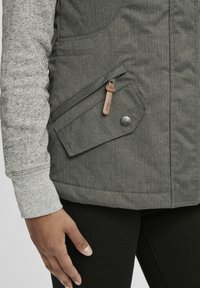 Grå pufferväst med en slät yta, utrustad med en stor sidoficka, knappdetaljer och en kontrasterande texturerad ärm i ljusgrått.
