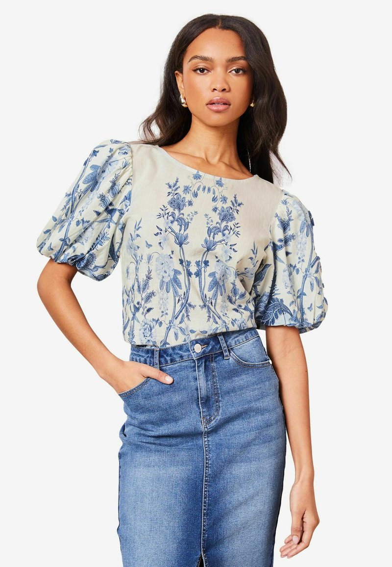 Bloemmotief blouse van lichte stof, met pofmouwen en een ronde halslijn. Draagt bij een blauwe denim rok en met de handen in de zakken.