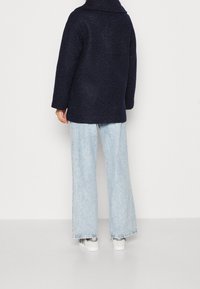 Merenvärinen teksturoitu takki, jossa on leveä kaulus, rento malli, yhdistettynä korkeavyötäröisiin vaaleansinisiin farkkuihin ja valkoisiin tennareihin, seisoo valkoista taustaa vasten.