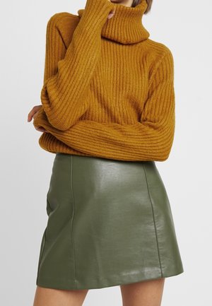 Femme portant un pull à col roulé côtelé moutarde et une jupe en cuir vert olive, les bras croisés avec une main touchant le visage.