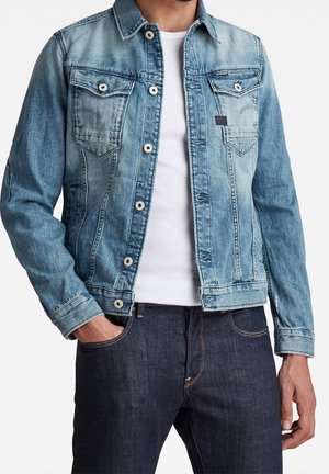 Homme portant une veste en jean bleu clair par-dessus un t-shirt blanc avec un jean bleu foncé, une main dans la poche, debout devant un fond blanc.