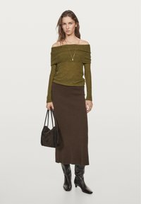 Grüner schulterfreier Ripp-Pullover, brauer taillierter Rock in Kniehöhe, schwarze kniehohe Stiefel und eine schwarze Handtasche. Einfaches silbernes Halsband sichtbar.
