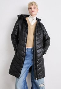 Svart quiltet pufferjakke med hette, med full glidelås, sidelommer og en glatt, shiny tekstur. Brukt over en beige genser og blå jeans.