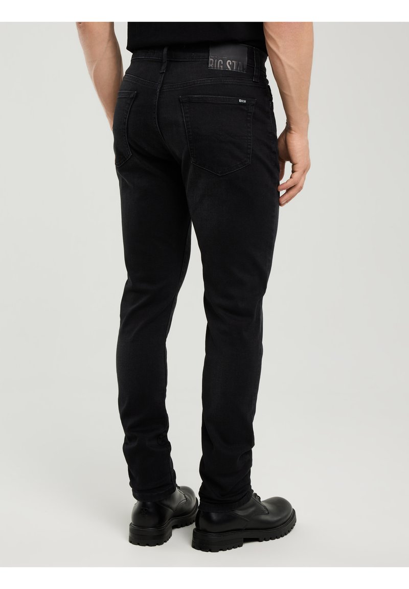 Bărbat purtând blugi negri slim-fit și cizme negre din piele, stând cu o mână relaxată pe lângă el, cu spatele către privitor.