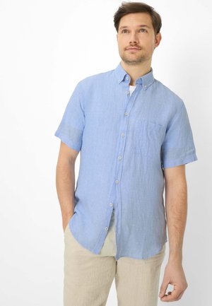 Mann trägt hellblaues kurzärmeliges Hemd mit Knöpfen und beigefarbene Hose, steht mit einer Hand in der Hosentasche vor weißem Hintergrund.