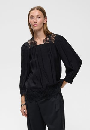 Mujer con cabello liso castaño que lleva una blusa negra con detalles de encaje en los hombros y el dobladillo, combinada con pantalones negros de pierna ancha.