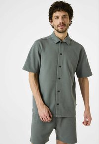 Camisa de botón de manga corta, con cuello, en una suave tela verde texturizada, combinada con pantalones cortos a juego, con botones negros y un diseño minimalista.