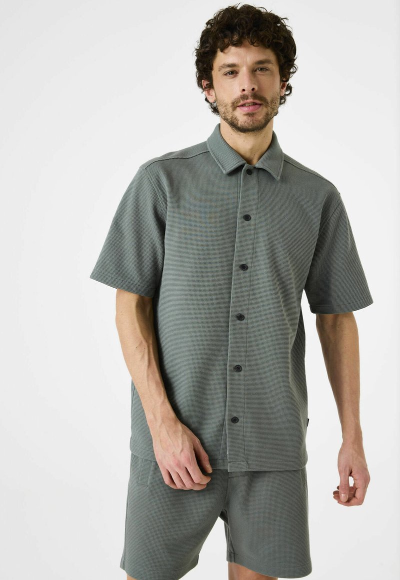 Camisa de botón de manga corta, con cuello, en una suave tela verde texturizada, combinada con pantalones cortos a juego, con botones negros y un diseño minimalista.
