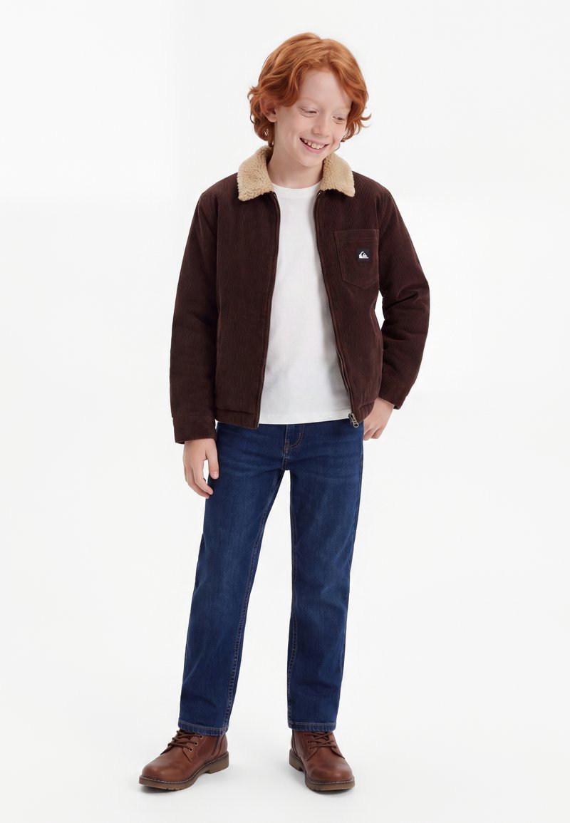 Garçon aux cheveux roux portant une veste marron avec un col beige, une chemise blanche, un jean bleu et des bottes marron, debout et souriant.