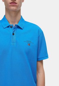 Blå pikétröja i texturerad tyg. Har en klassisk krage, tv å svarta knappar och en liten broderad logotyp med en flagga.