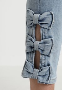 Ljusblå denim capri byxor med tre dekorativa tygrosetter längs sidsömmarna, med synliga sömmar.