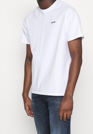 Homme portant un t-shirt Levi's blanc uni et un jean bleu, debout devant un fond blanc avec les bras détendus le long du corps.