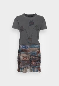 Grå bomullströja med en grafisk teapot och text ovanför, kombinerad med en rynkad kjol som har blandade mönstrade tyger i olika färger och texturer.