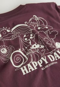 T-shirt bordeaux avec un design graphique blanc représentant des personnages souriants, y compris un escargot, des fleurs, des champignons et des arbres, avec le texte "JOUR HEUREUX".