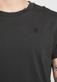T-shirt in cotone grigio scuro con colletto rotondo, caratterizzato da un logo nero discreto sul lato sinistro del petto. Morbido al tatto, maniche corte e vestibilità rilassata.