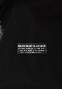 Etiqueta de tela negra con texto blanco que detalla la marca y el eslogan. Los bordes están cosidos y la etiqueta tiene forma rectangular.