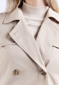 Manteau beige à double boutonnage avec de gros boutons, col et poches latérales. Texture douce avec une finition lisse, superposé à un col roulé clair.