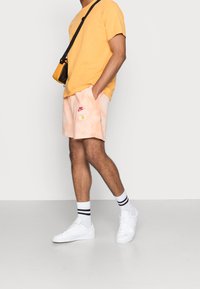 Pantalones cortos tie-dye en color durazno con un diseño de margaritas blancas y logo de Nike en rojo, combinados con una camiseta amarilla y zapatillas blancas con rayas negras.