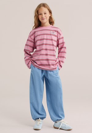 Jeune fille portant un t-shirt à manches longues rayé rose, un jean ample bleu clair et des baskets bleu clair, debout les mains dans les poches.