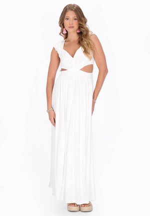 Femeie cu păr lung și ondulat, purtând o rochie maxi albă fără mâneci, cu decupaje laterale și cercei roz cu ciucuri, stând în fața unui fundal alb.