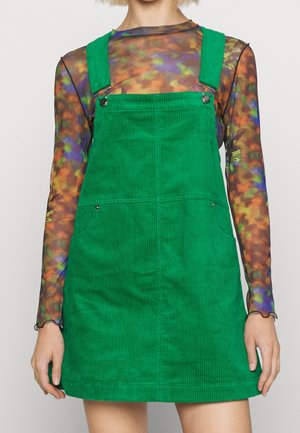 Frau trägt ein leuchtend grünes Kord-Latzkleid über einem bunten, langärmeligen, durchsichtigen Mesh-Oberteil mit einem mehrfarbigen abstrakten Muster.