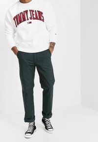 Weißer Pullover mit roter Schrift "TOMMY JEANS", kombiniert mit dunkelgrünen Hosen und schwarzen High-Top Sneakers, vor einem hellen Hintergrund.