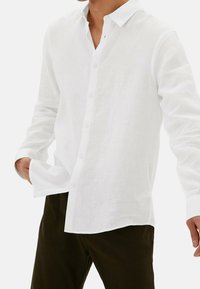 Camisa de lino blanca con un diseño abotonado, que presenta un cuello y un corte relajado. Mangas largas y un pequeño detalle en forma de lazo en el cuello.