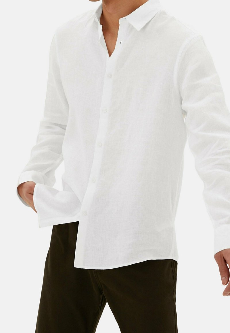 Camisa de lino blanca con un diseño abotonado, que presenta un cuello y un corte relajado. Mangas largas y un pequeño detalle en forma de lazo en el cuello.