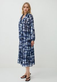 Donna che indossa una blouse a maniche lunghe a volant blu e bianca a scacchi, abbinata a una gonna midi plissettata, con sandali neri con tacco.