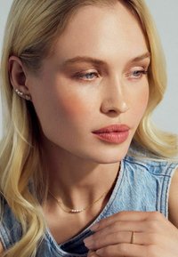 Guldhalsband med pärlor, guldring och flera örhängen. Modellen har rakt blont hår, ljus hy och en denimtop. Naturlig sminkning.